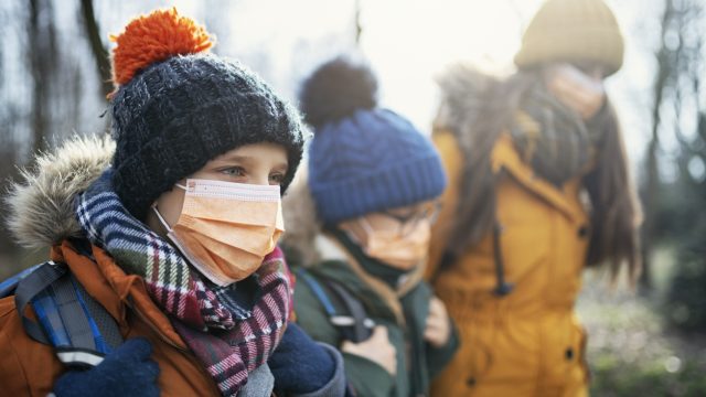 Three kids wearing back packs, face masks, and winter clothing walk outdoors.