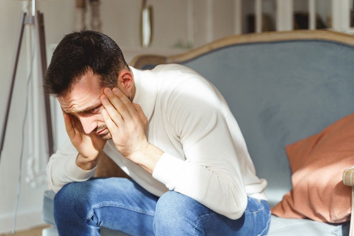 Maschio triste in un pullover bianco che piega il doppio con un dolore su un divano