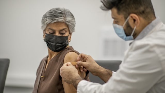 A male doctor puts a band aid on a senior woman's arm after he administered the COVID-19 vaccine injection. They are both wearing a protective face mask to protect themselves from the transfer of germs.