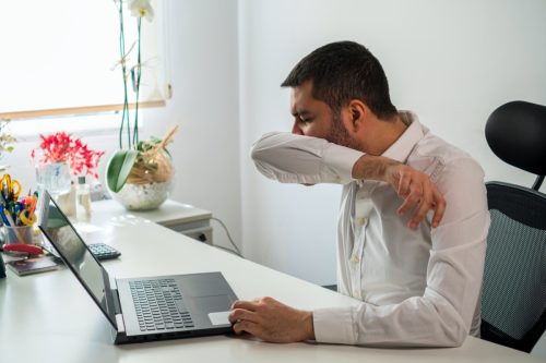 man coughing into arm at desk in front of laptop