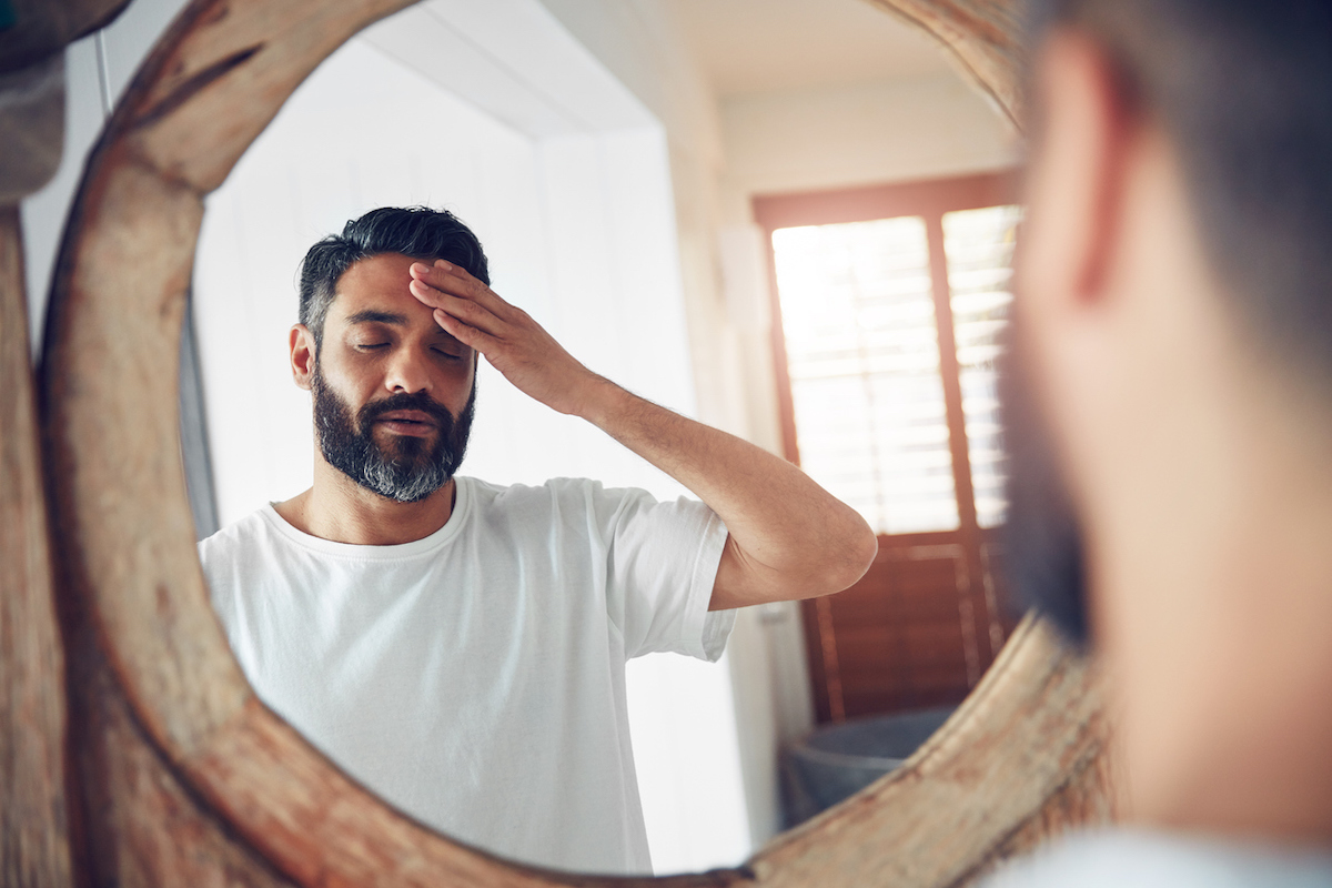 Shot of a man standing in front of the bathroom mirror looking exhausted, holding his head