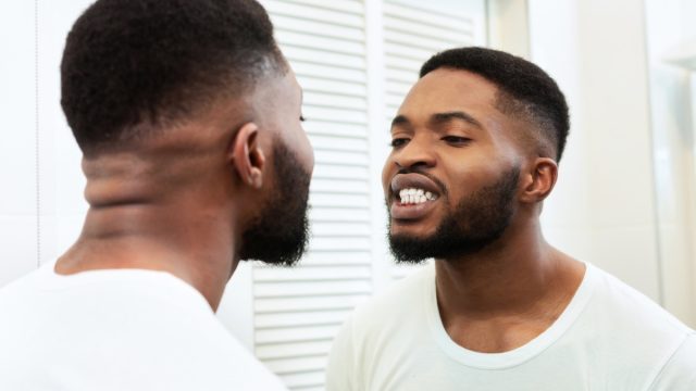 Young man looking in mirror at his mouth and teeth in bathroom, checking their health state