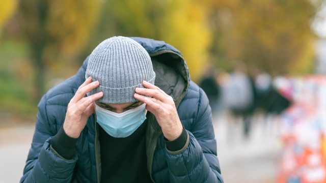 A man in a medical mask in the park with his hand on his head due to a headache.