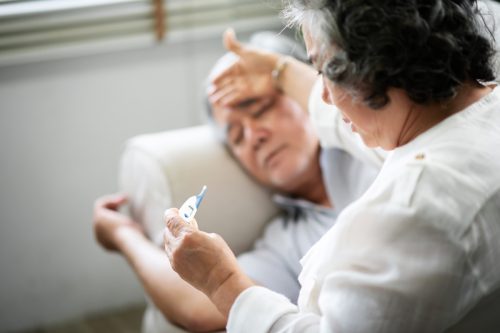 Un bătrân bolnav stă întins pe canapea în timp ce soția lui ține termometrul și se uită la el