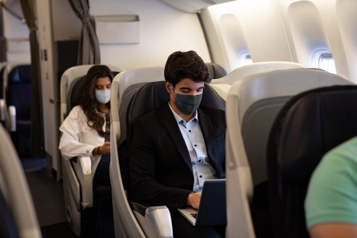 Business man traveling and wearing a facemask on the plane while using his laptop– COVID-19 pandemic lifestyle concepts