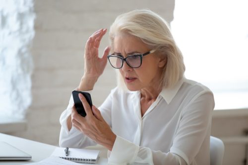 A senior woman wearing glasses and sitting at a desk looks at her smartphone with a surprised and confused look, maybe the victim of a scam.