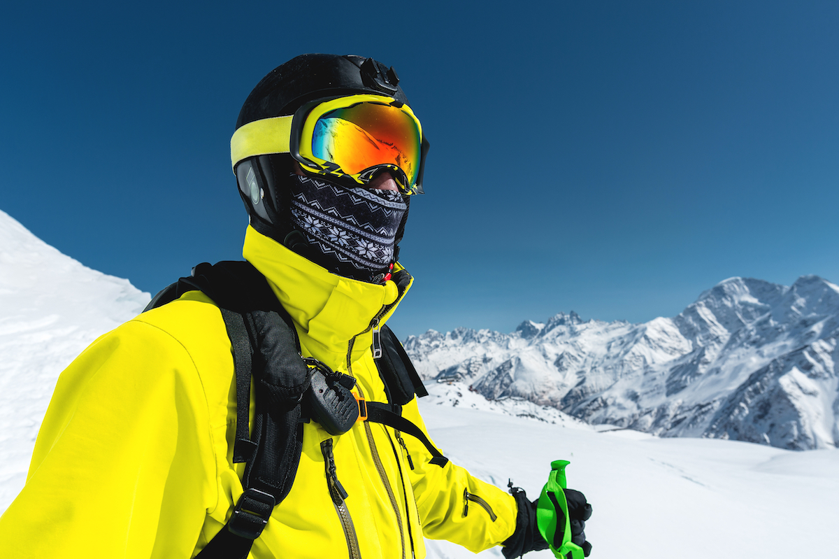 Close-up portrait of a skier in a mask and helmet with a closed face against a background of snow-capped mountains and blue sky