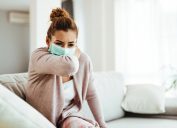 Woman wearing sneezing into her elbow while wearing protective face mask at home.