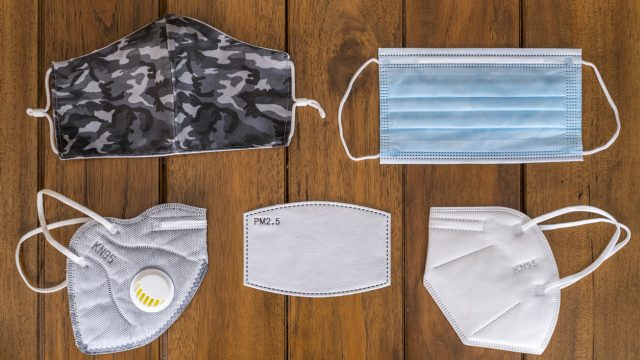 Five different types of face masks and filters resting on a wooden background, including a cloth mask, KN95, and surgical mask