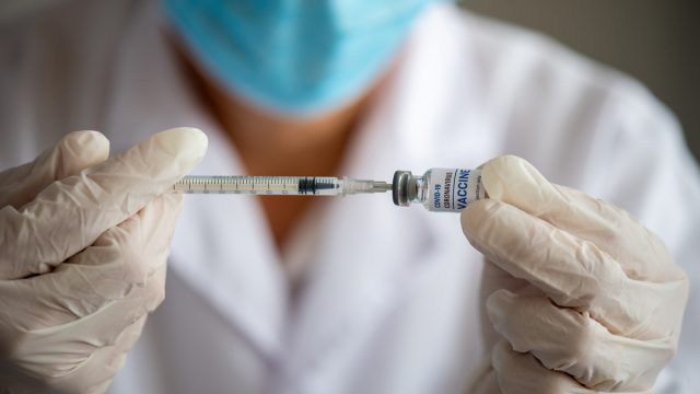 Hand with white surgical gloves taking coronavirus vaccine dose from vial with syringe