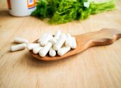White vitamin supplement capsules sitting in a wooden spoon on a table top in front of green herbs.