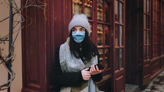 Woman in a coat, a scarf, a hat and a protective medical blue mask is standing in street