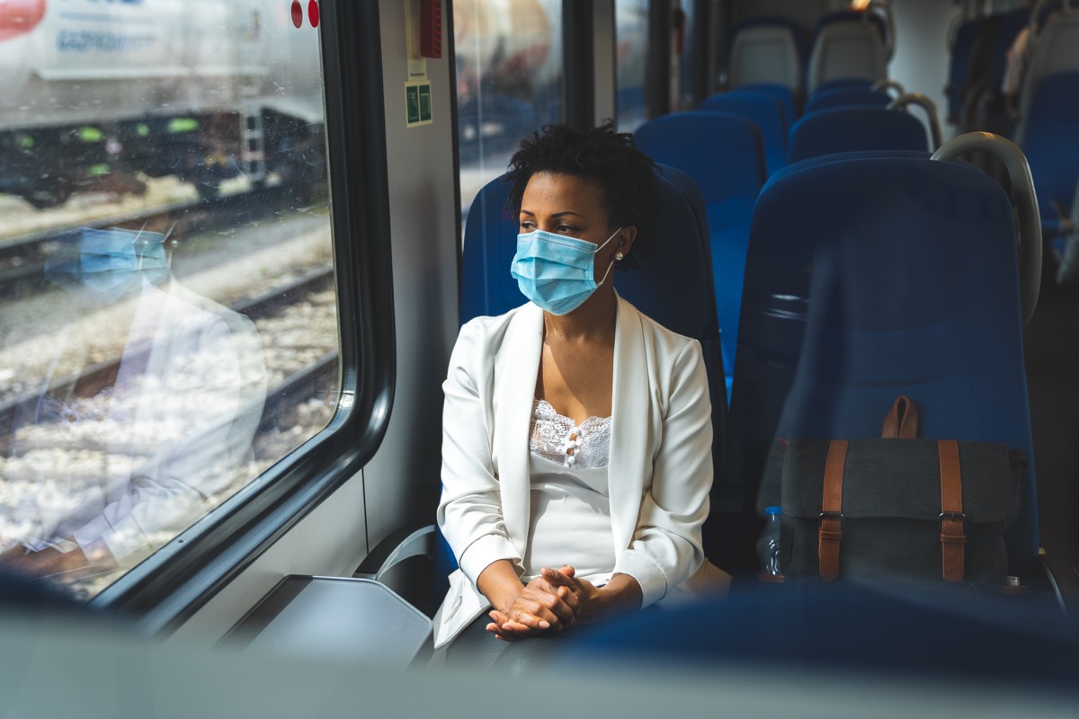 Woman sitting in the train and wearing a protective mask. She is looking trough the window.