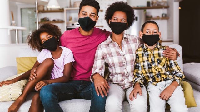A young family of four wearing face masks while sitting on their couch.