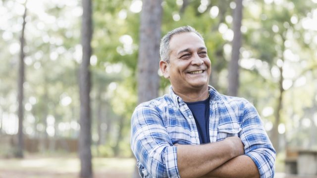 A mature man in his 50s wearing a plaid shirt, standing in a park, smiling with his arms crossed.