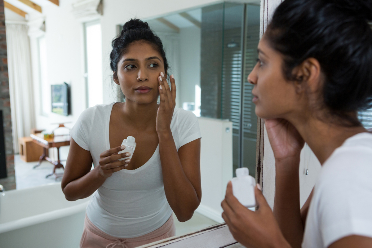 Young woman applying lotion or sunscreen while looking in mirror in bathroom