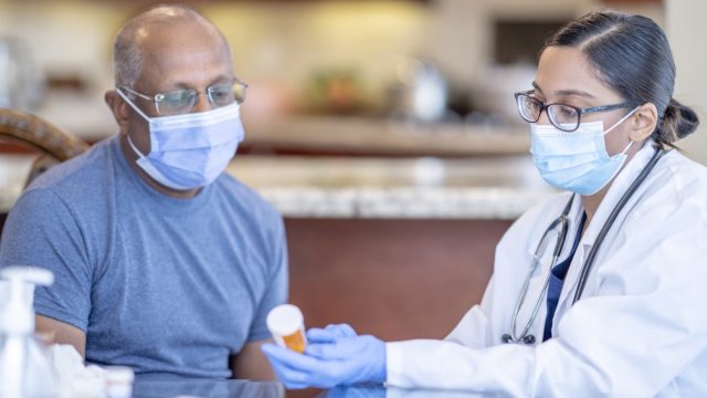 A female doctor is holding a prescription bottle that belongs to her patient. She is reviewing the medicinal ingredients as his patient is seated next to her. Both patient and doctor is using a face mask to help prevent the spread of germs.