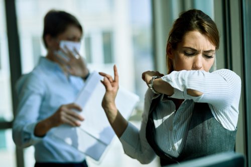 Young businesswoman coughing into elbow in the office. Her coworker is in the background.