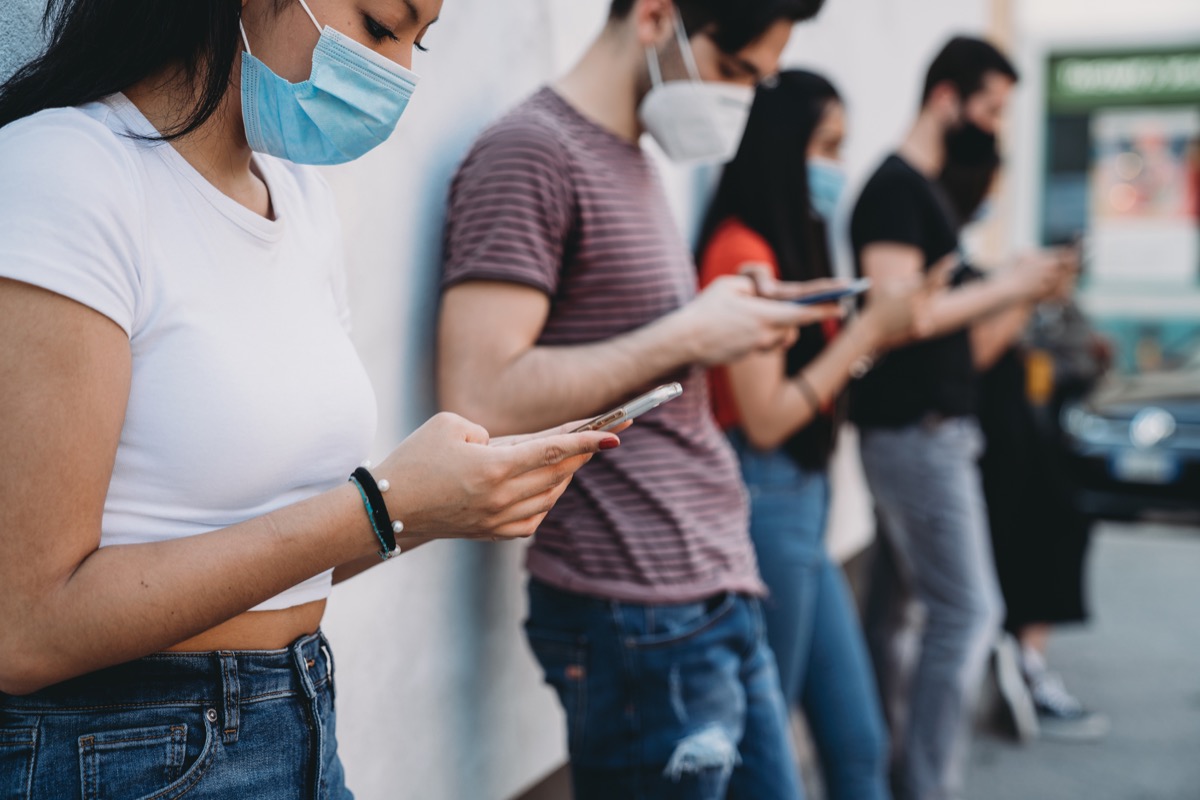 Eine Gruppe junger Freunde steht vor einer Wand, benutzt Smartphones und trägt Schutzmasken.