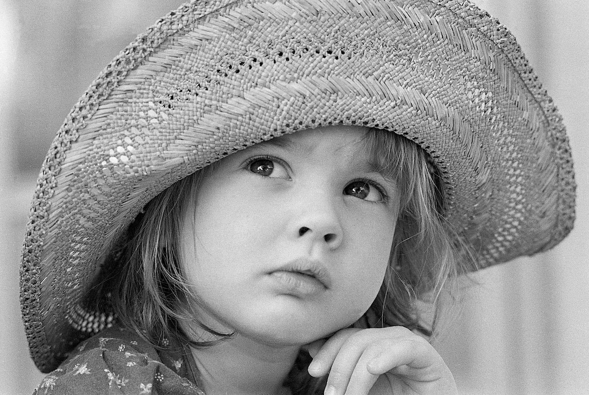 Four year old Drew Barrymore, the youngest member of the Barrymore and Drew families, looks upward wearing a straw hat.