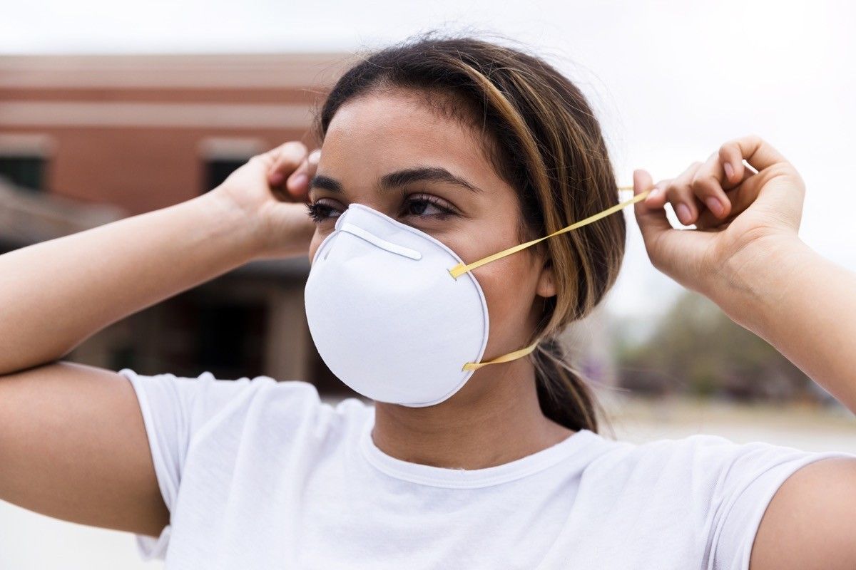 During a pandemic, a mid adult woman protects herself by placing an N95 face mask over her nose and mouth. She is standing outdoors.