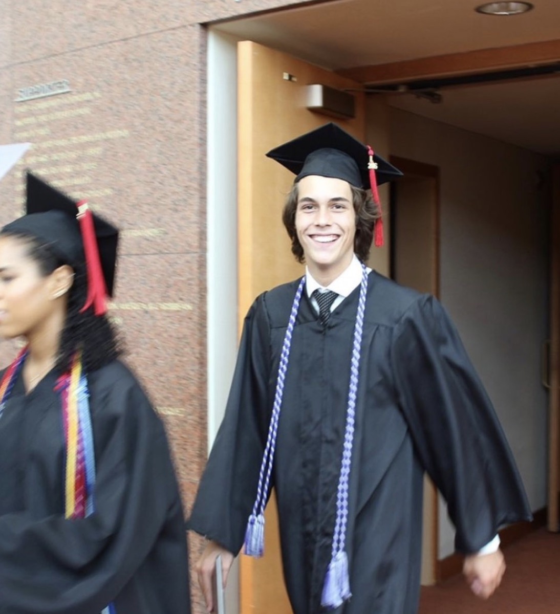 flynn busson in cap and gown emerging from a brick building