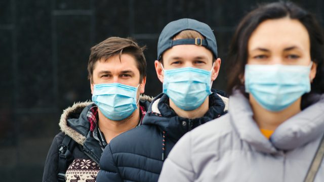 A group of three young people wearing face masks and winter coats stand in a line.