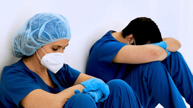 A male and female healthcare worker wearing face masks and gloves sit against a wall while looking tired and overworked.