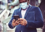 A man wearing a blue blazer and protective face mask checks his smartphone.