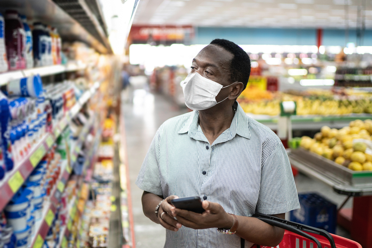 Mature man in mask using mobile phone and choosing products in supermarket, potentially for Super Bowl party