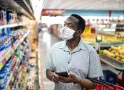 Mature man in mask using mobile phone and choosing products in supermarket, potentially for Super Bowl party
