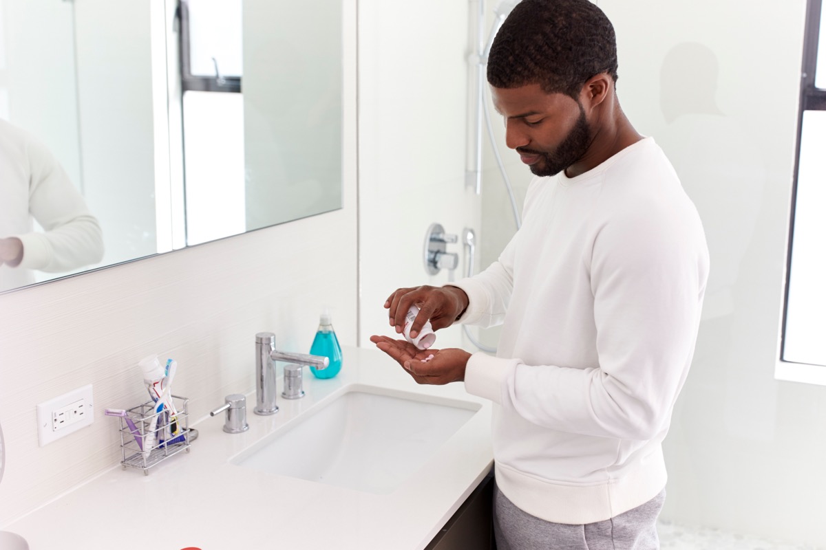 young man in white shirt standing in white bathroom taking vitamins out of a pill bottle in his hand