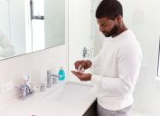 young man in white shirt standing in white bathroom taking vitamins out of a pill bottle in his hand