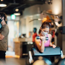 Two sports persons are wearing protective face masks and training in a gym while keeping social distancing.