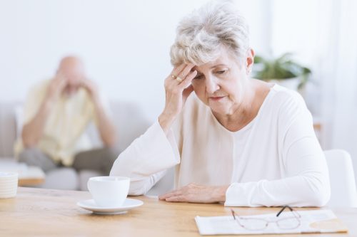 A senior woman sits at a table in front of a coffee while holding her head with a distressed look on her face
