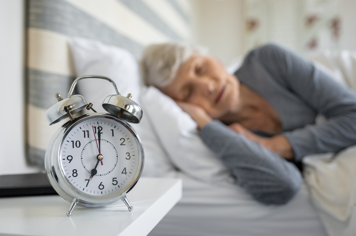 Крупен план на будилник със старша жена в дълбок сън на заден план