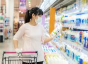 Woman with hygienic mask and rubber gloves with shopping cart in grocery store looking for daily fresh milk to buy during covid-19 outbreak for preparation for a pandemic quarantine