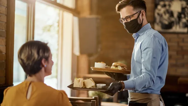 woman being served in a restaurant by young waiter wearing black mask