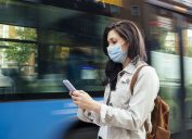 A young woman wearing a face mask checks her smartphone while waiting for a city bus