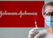 A woman wearing protective glasses, a face mask, and a hair covering holds a syringe in front of a Johnson & Johnson sign