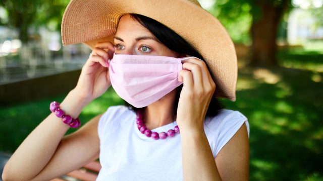 A young woman wearing a large straw hat puts on a pink face mask while standing outdoors