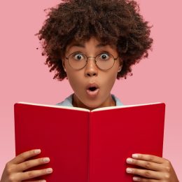 Smart young woman with glasses looking amazed while reading a book