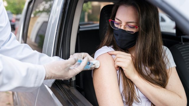 A young woman sitting in a car and wearing a face mask receives the COVID-19 vaccine from a healthcare worker wearing gloves.