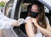 A young woman sitting in a car and wearing a face mask receives the COVID-19 vaccine from a healthcare worker wearing gloves.