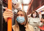A young woman riding a subway train while wearing a mask, standing in front of another woman who is checking her smartphone