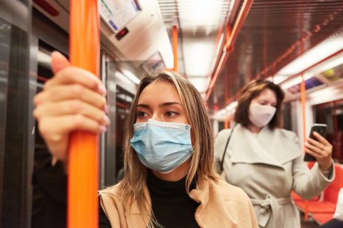 A young woman riding a subway train while wearing a mask, standing in front of another woman who is checking her smartphone