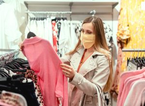 A young woman wearing a face mask picks up a shirt while shopping in a department store.
