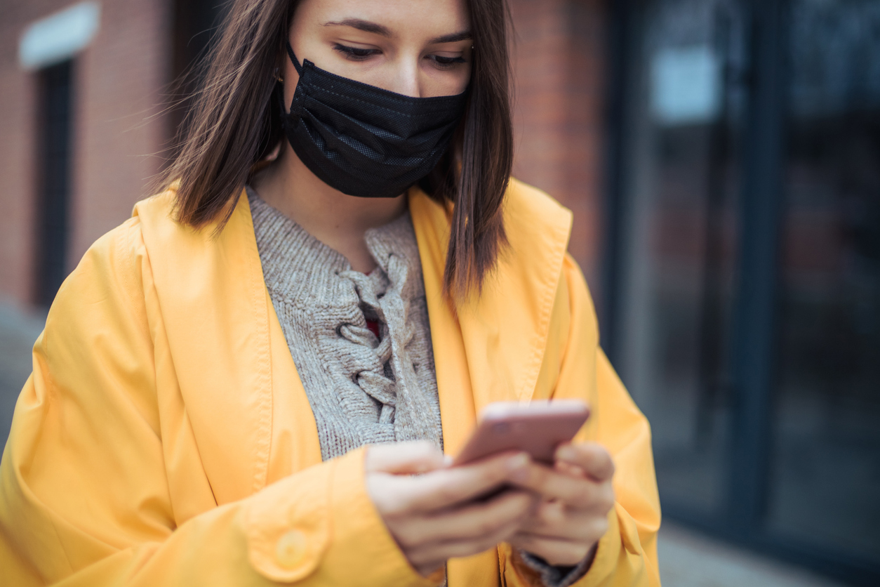 A young woman wearing a yellow jacket and a face mask checks her smartphone.