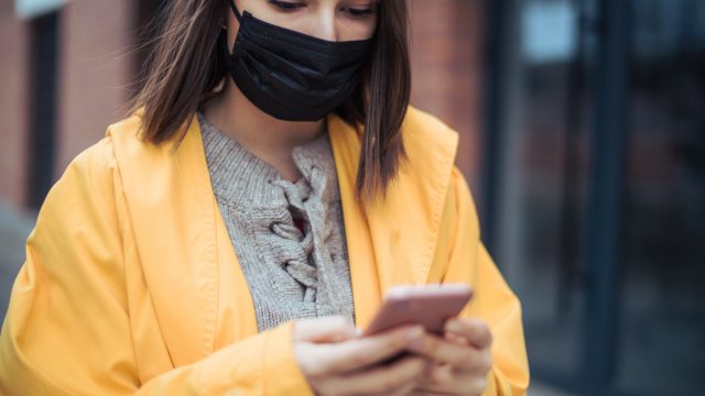 A young woman wearing a yellow jacket and a face mask checks her smartphone.