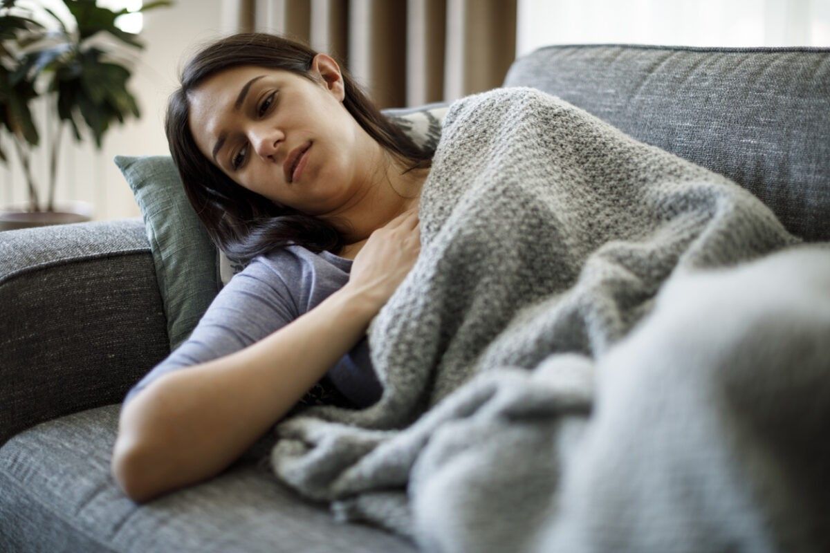 A young woman is lying on the sofa as she is covered with a blanket and a tired expression on her face